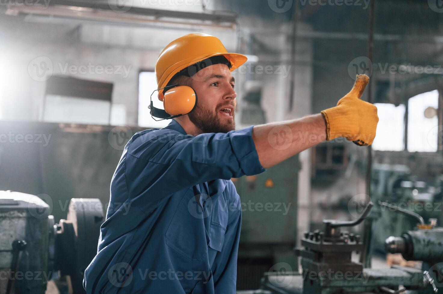 Man in hard hat and ,headphones is showing thumb up. Factory worker in blue uniform is indoors photo