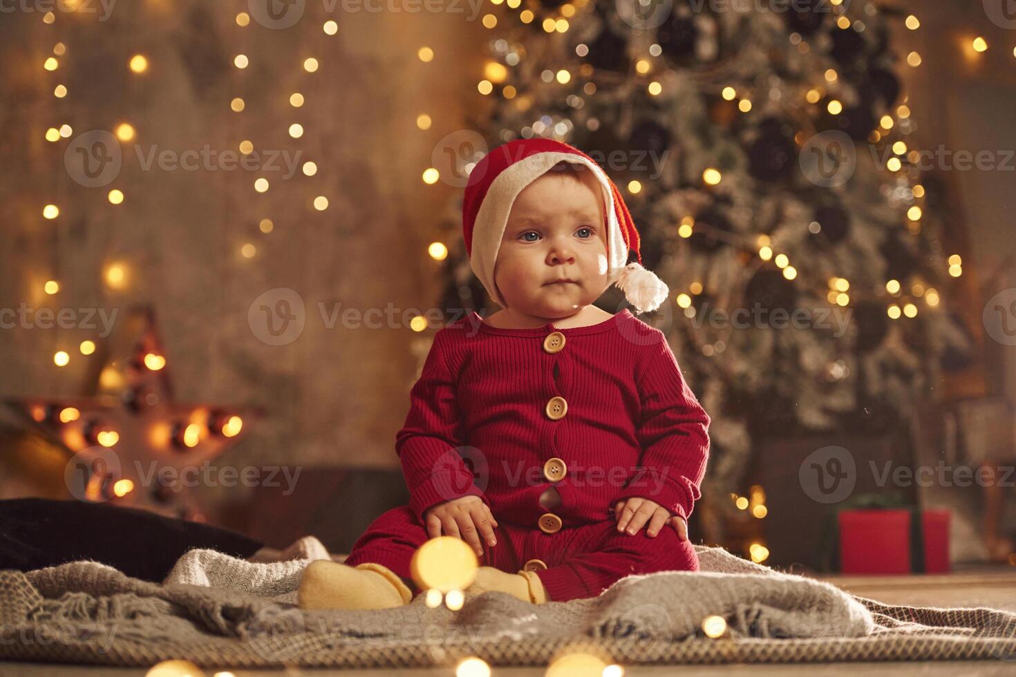 Sitting ,in festive clothes and Santa hat. Cute little baby is at home. New year decorations. Conception of holidays photo