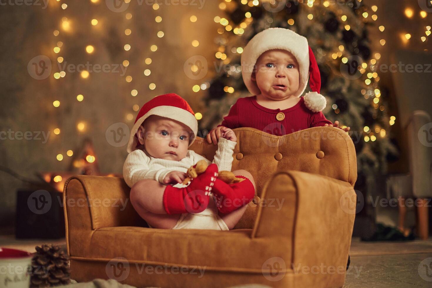 Baby is on the little sofa. .Two, kids in Santa hats are celebrating holidays indoors together photo