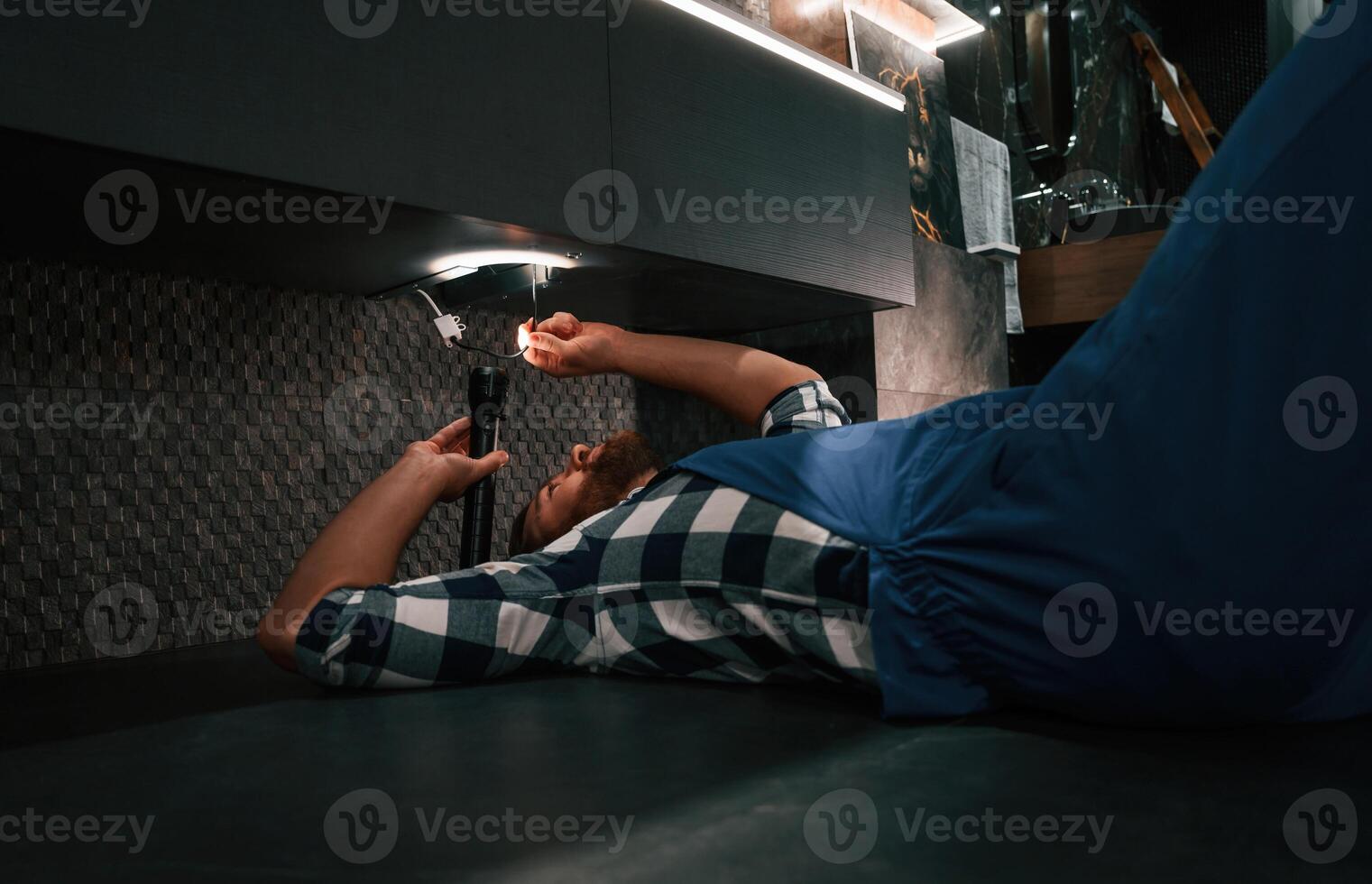 Using flashlight.. Fixing water tap. Plumber in blue uniform is at work in the bathroom photo