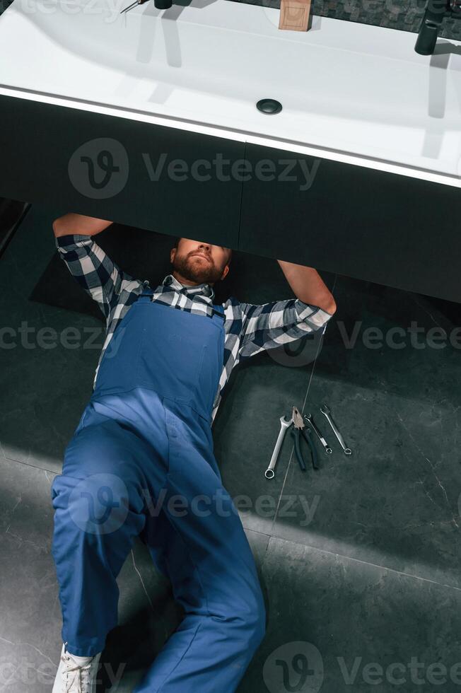 Lying down, on the floor, fixing the siphon. Plumber in blue uniform is at work in the bathroom photo