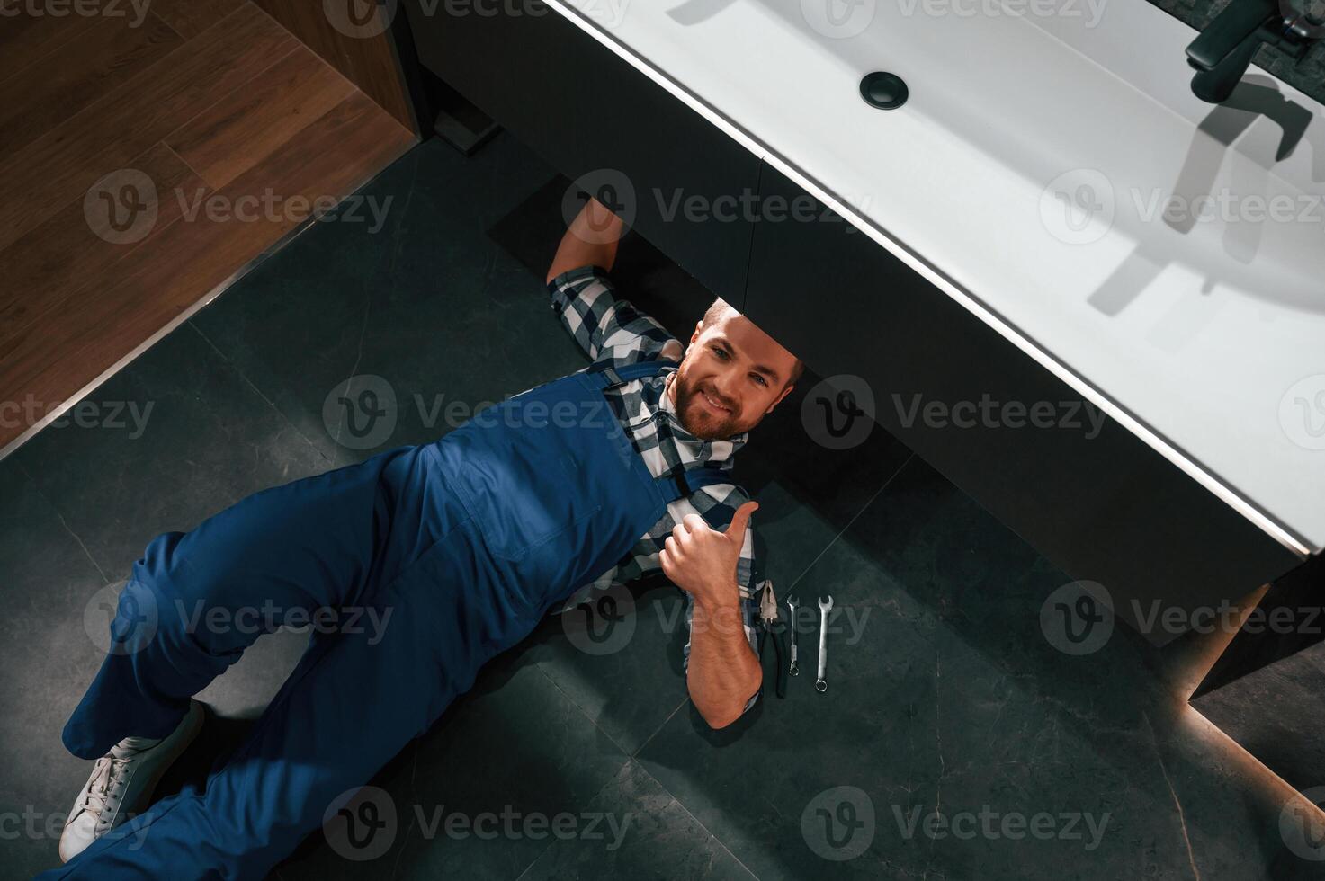 Lying, down on the floor, fixing the siphon. Plumber in blue uniform is at work in the bathroom photo