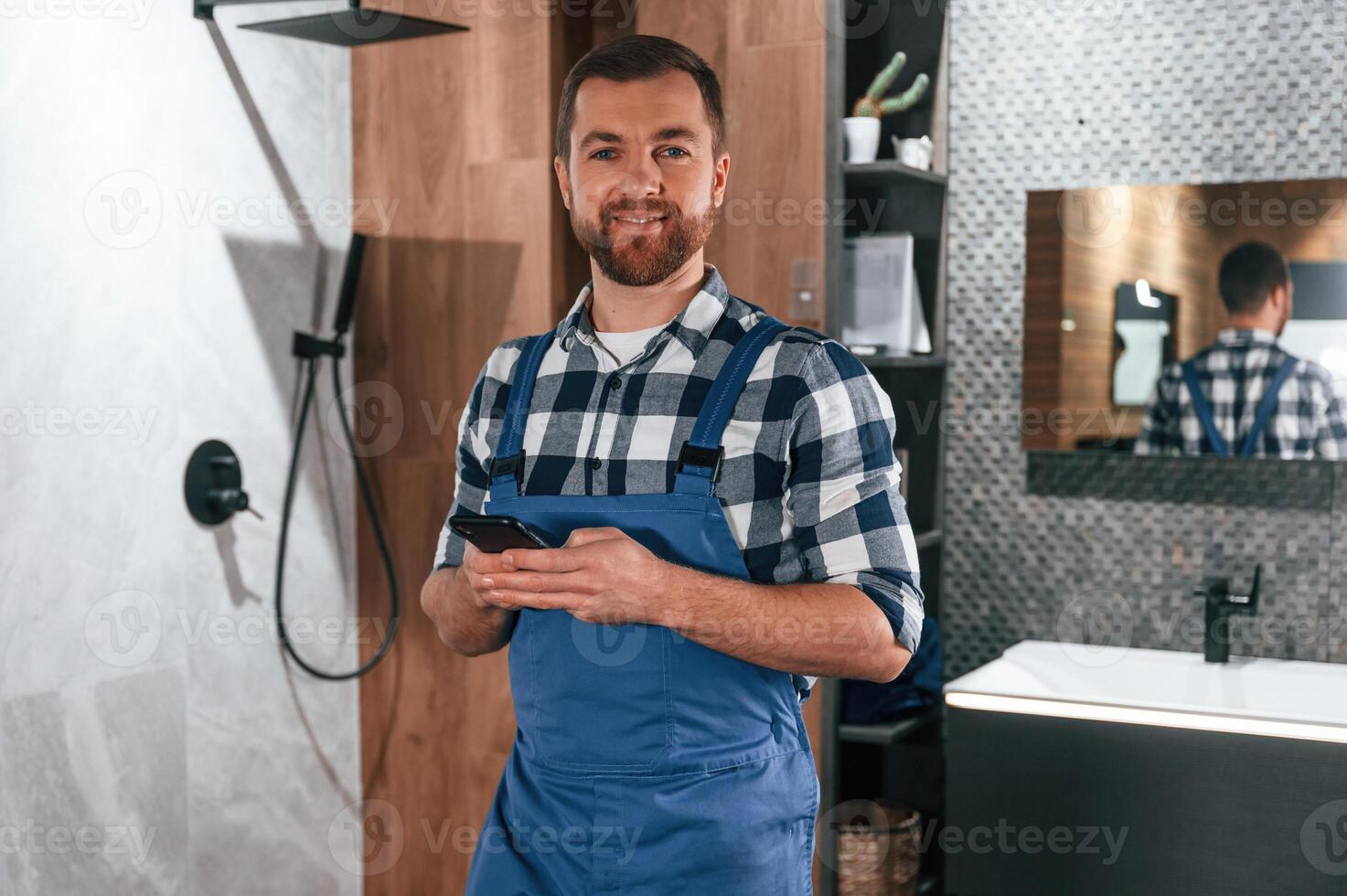 Standing and, holding smartphone. Plumber in blue uniform is at work in the bathroom photo