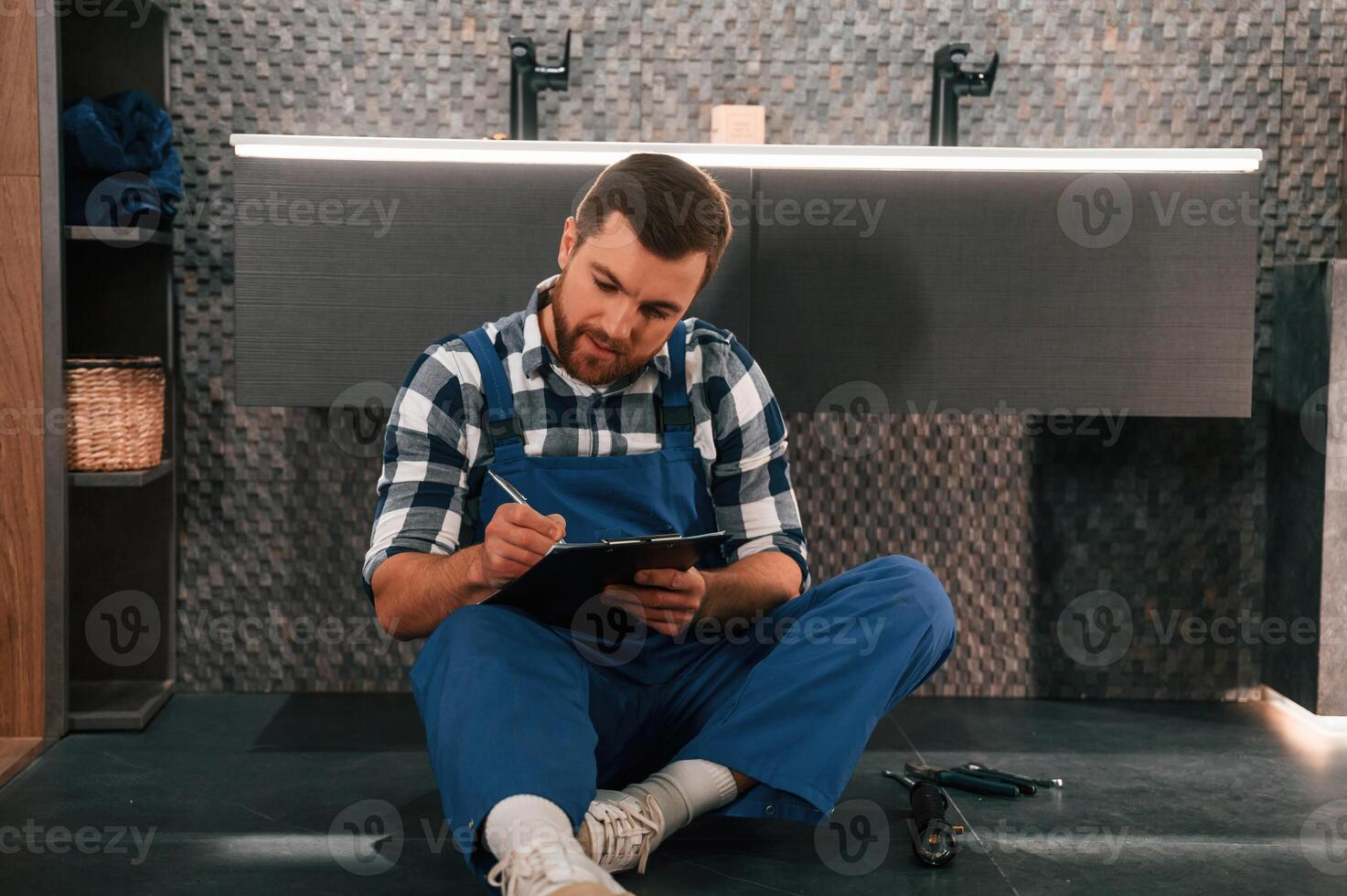 Sitting ,on the floor with notepad in hands. Plumber in blue uniform is at work in the bathroom photo