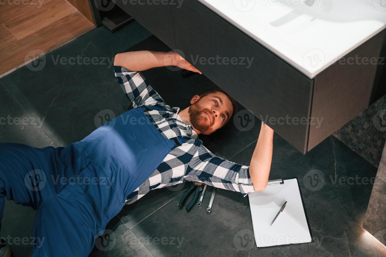 Fixing the siphone. Lying down on the floor. Plumber in blue uniform is at work in the bathroom photo