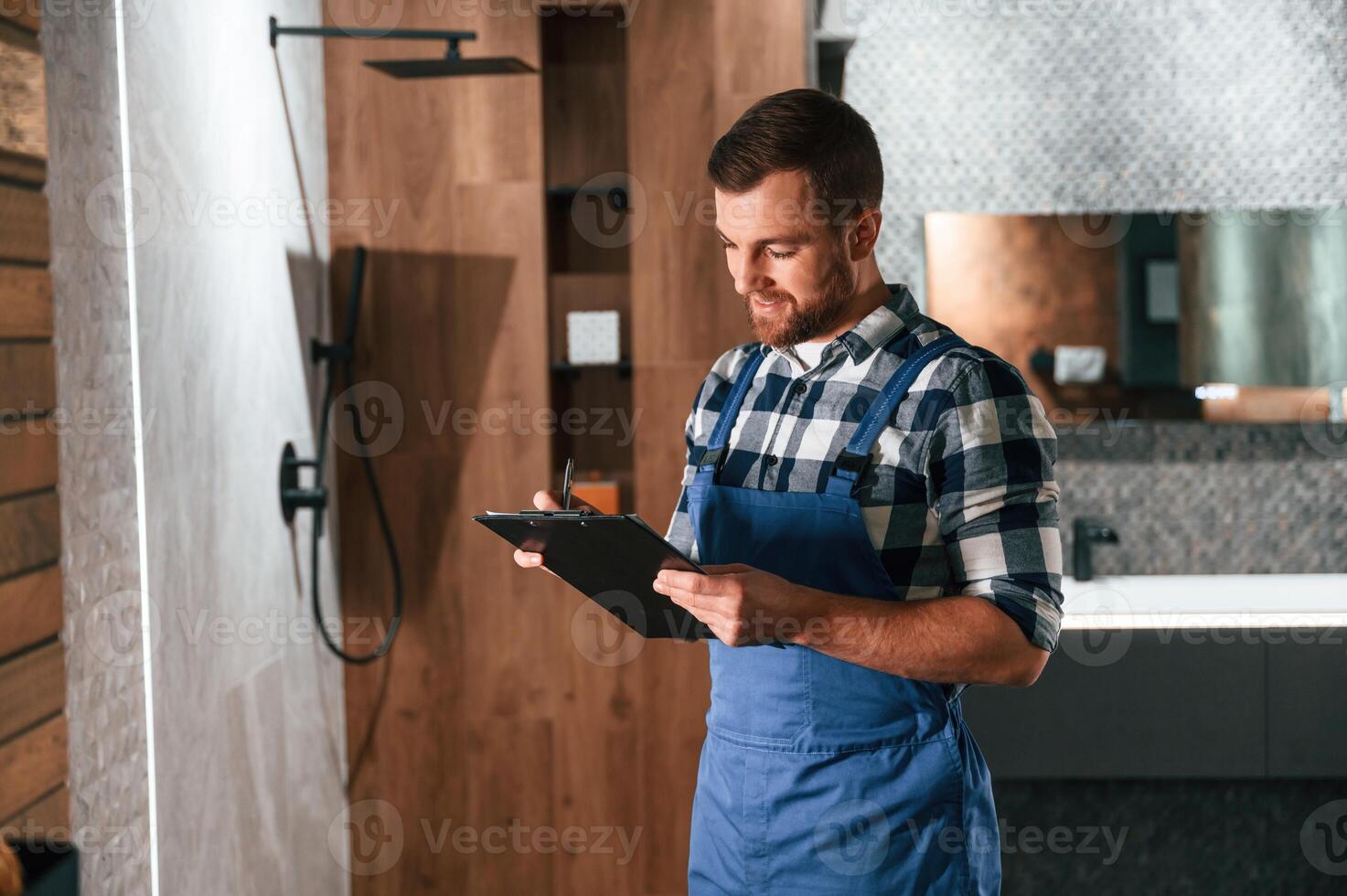 Standing and ,holding notepad. Plumber in blue uniform is at work in the bathroom photo