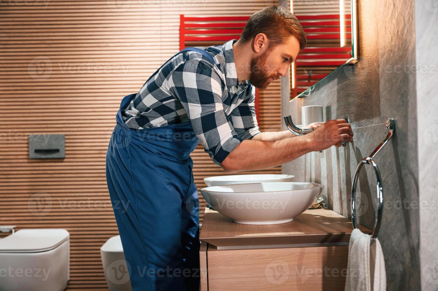 Uses a screwdriver for repairs.. Plumber, in blue uniform is at work in the bathroom photo