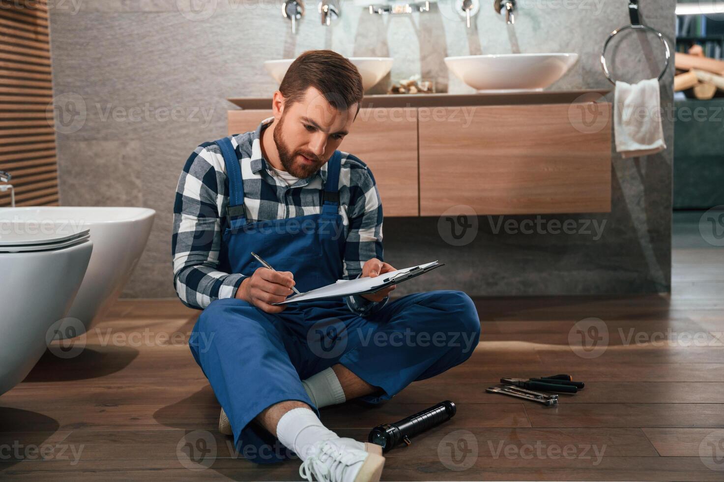 With notepad in hands. Plumber in blue uniform is at work in the bathroom photo