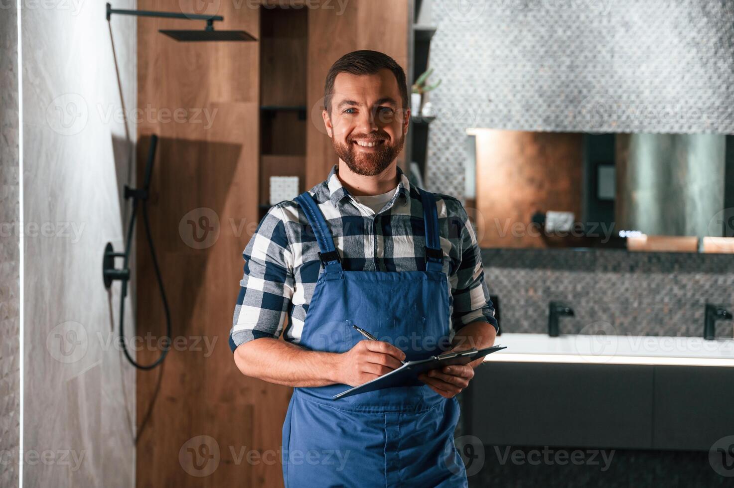Standing and holding notepad. .Plumber in blue uniform is at work in the bathroom photo