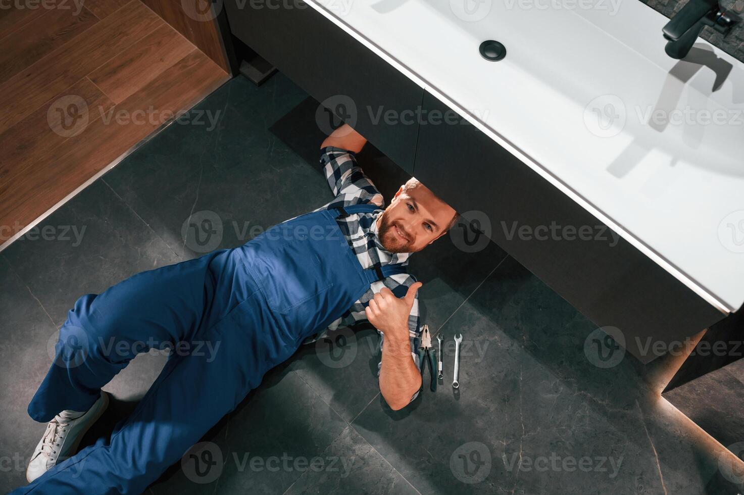 Lying down on the floor, fixing the siphon.. Plumber in blue uniform is at work in the bathroom photo