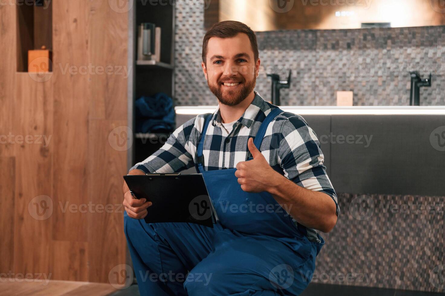Sitting on the floor with notepad in hands. Plumber, in blue uniform is at work in the bathroom photo