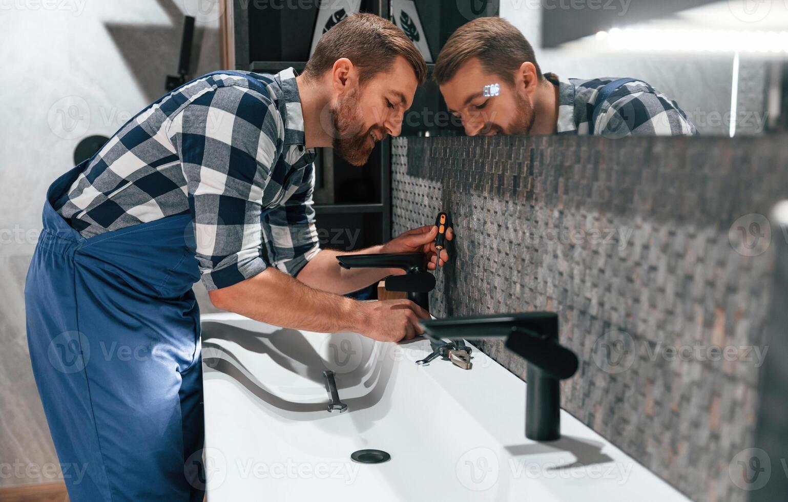 Side view.. Fixing, water tap. Plumber in blue uniform is at work in the bathroom photo