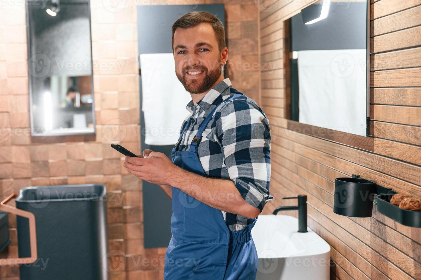 Smiling, holding smartphone in hands. Plumber in blue uniform is at work in the bathroom photo