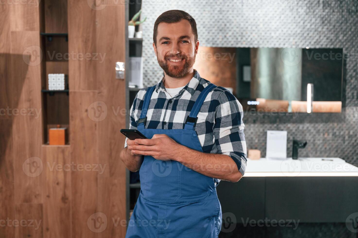 Standing ,and holding smartphone. Plumber in blue uniform is at work in the bathroom photo