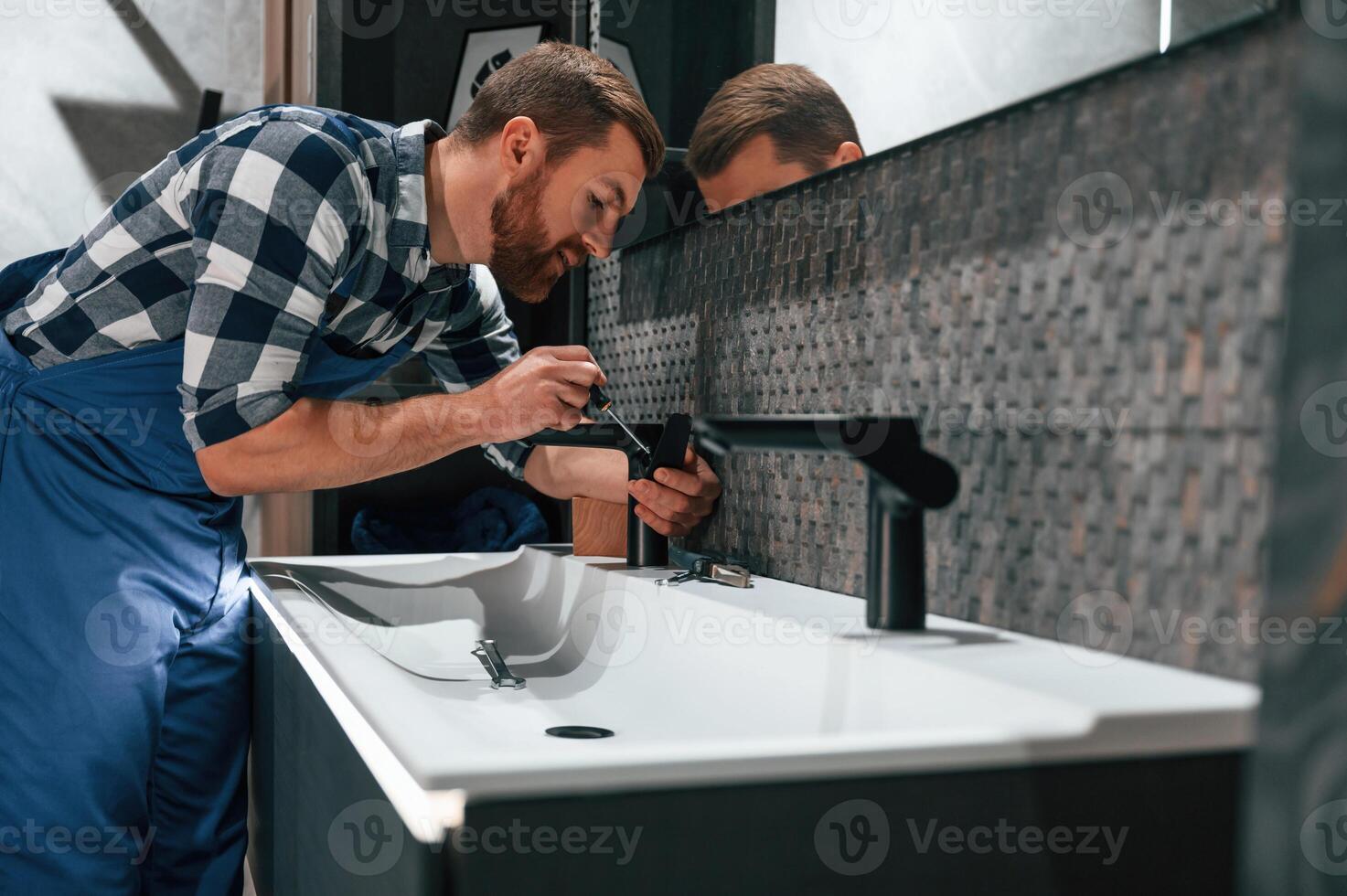 Side view.. Fixing water tap. Plumber in blue uniform is at work in the bathroom photo