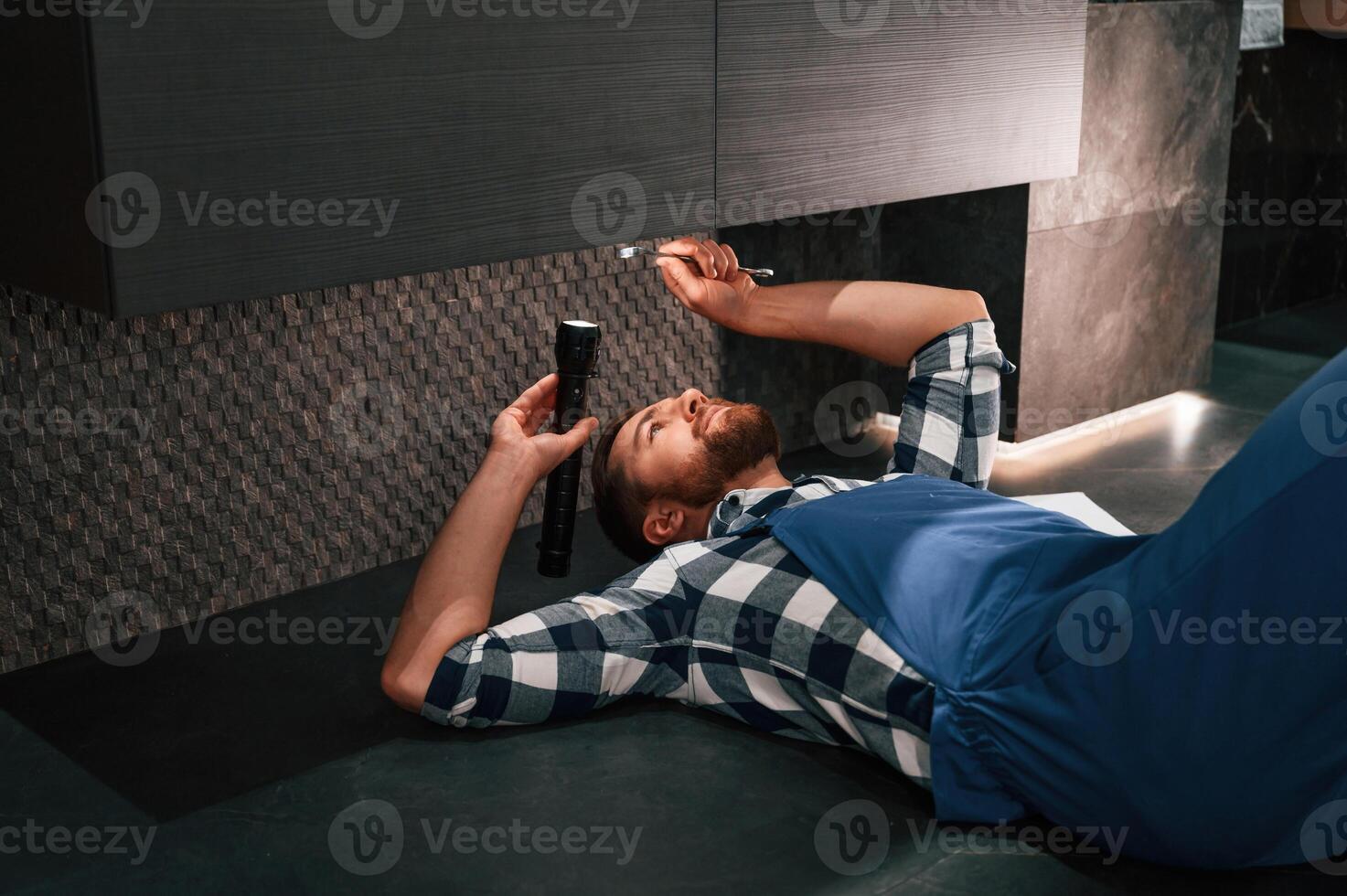 Fixing,, the siphone. Lying down on the floor. Plumber in blue uniform is at work in the bathroom photo