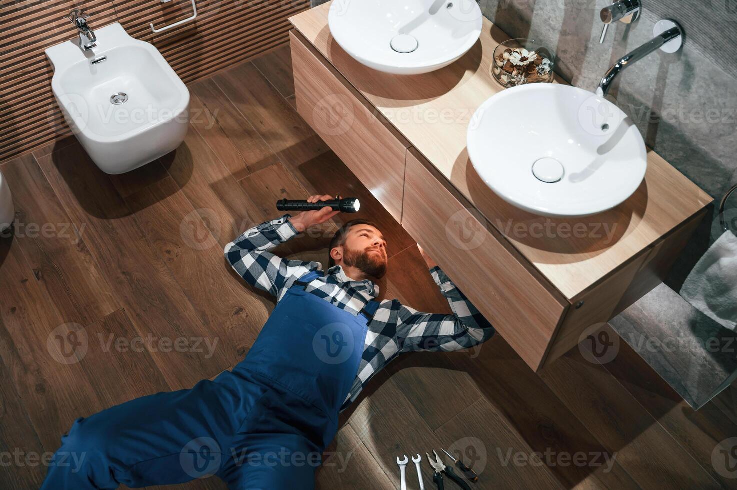 Top view. Lying down on the ground, fixing water, tap. Plumber in blue uniform is at work in the bathroom photo