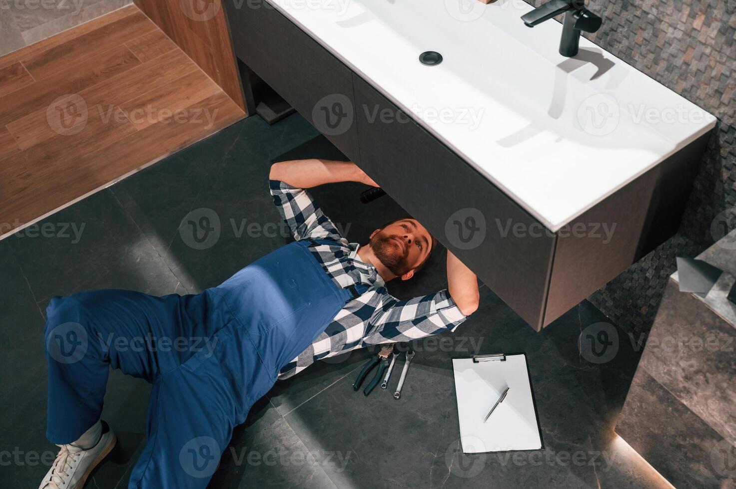 Fixing the siphone.. Lying down on the floor. Plumber in blue uniform is at work in the bathroom photo