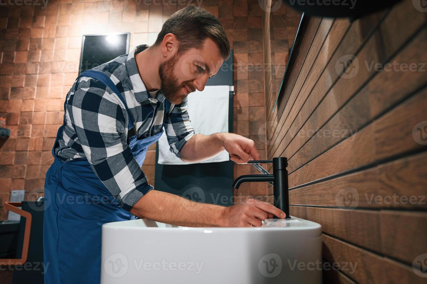 Standing by sink, and fixing water tap. Plumber in blue uniform is at work in the bathroom photo