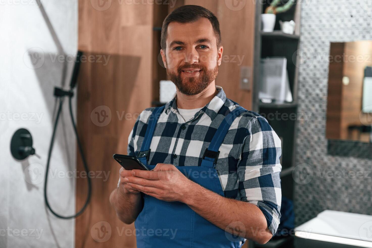 Standing and holding smartphone.. Plumber in blue uniform is at work in the bathroom photo