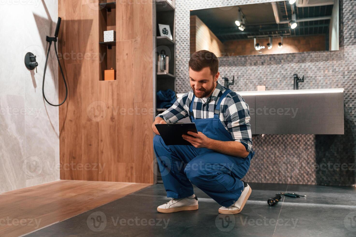 Sitting on the floor with notepad in hands. Plumber in blue uniform is at work in the bathroom photo