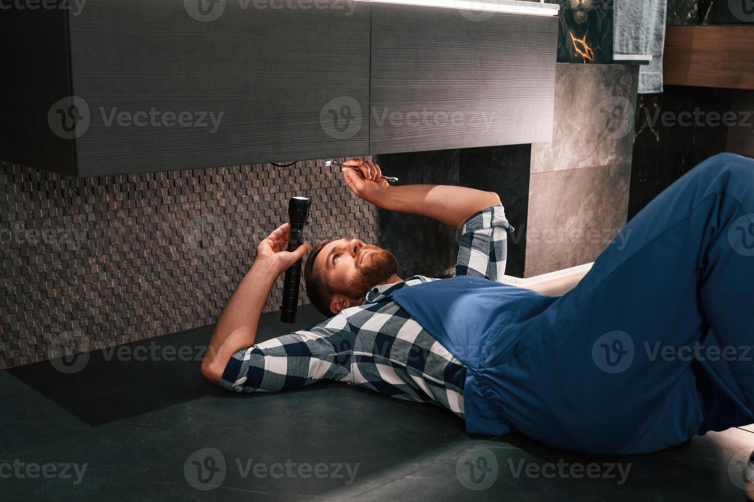 Using flashlight. Fixing water tap. Plumber in blue uniform is at work in the bathroom photo
