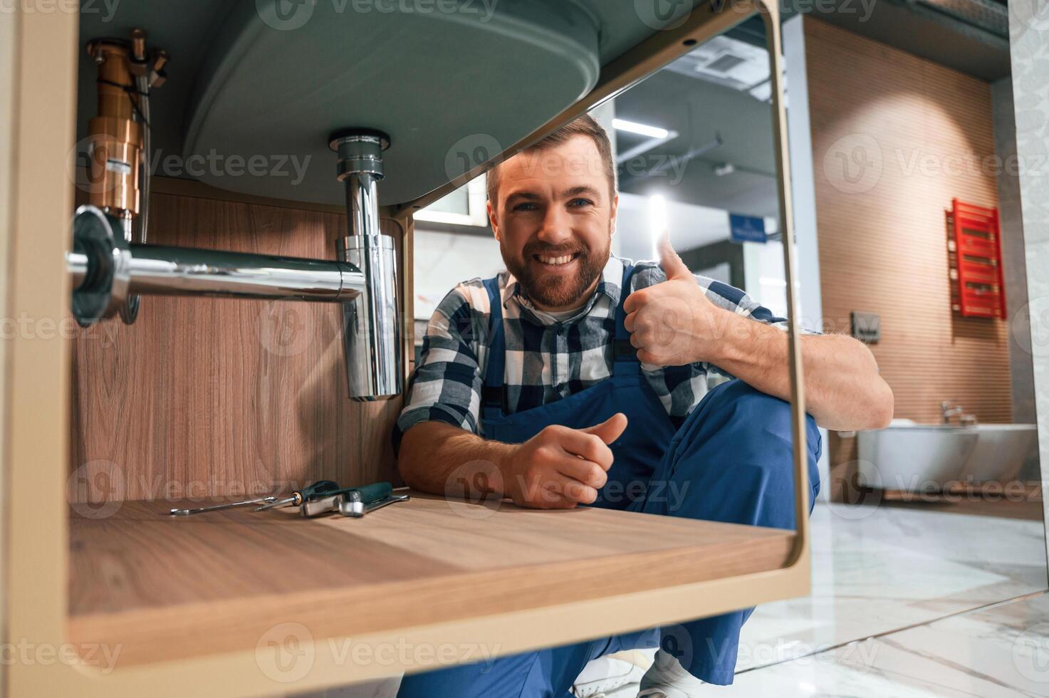 Shwoing thumb up and smiling. Plumber in blue uniform is at work in the bathroom photo