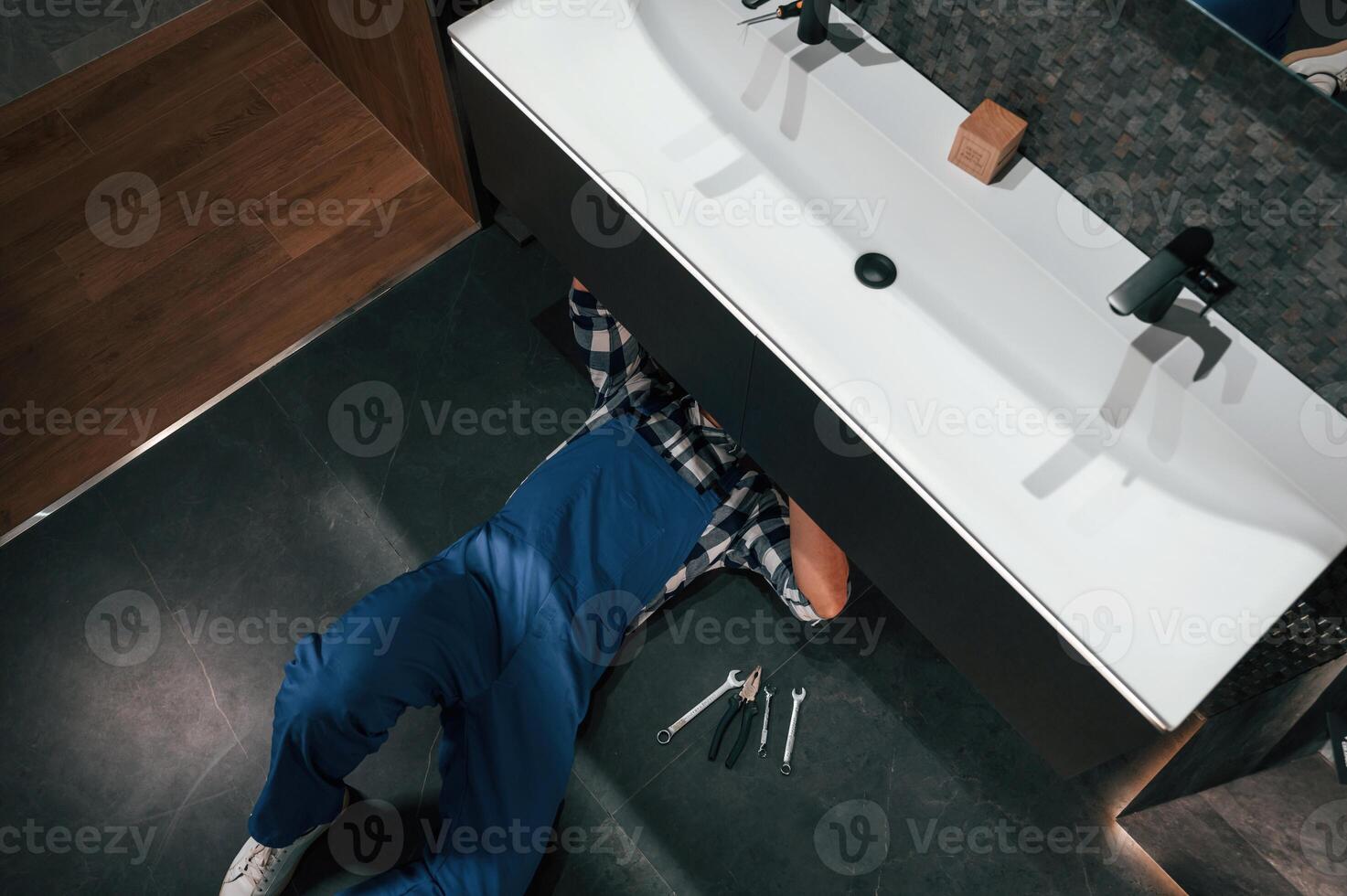 Lying down on the floor, fixing the siphon. Plumber in blue uniform is at work in the bathroom photo