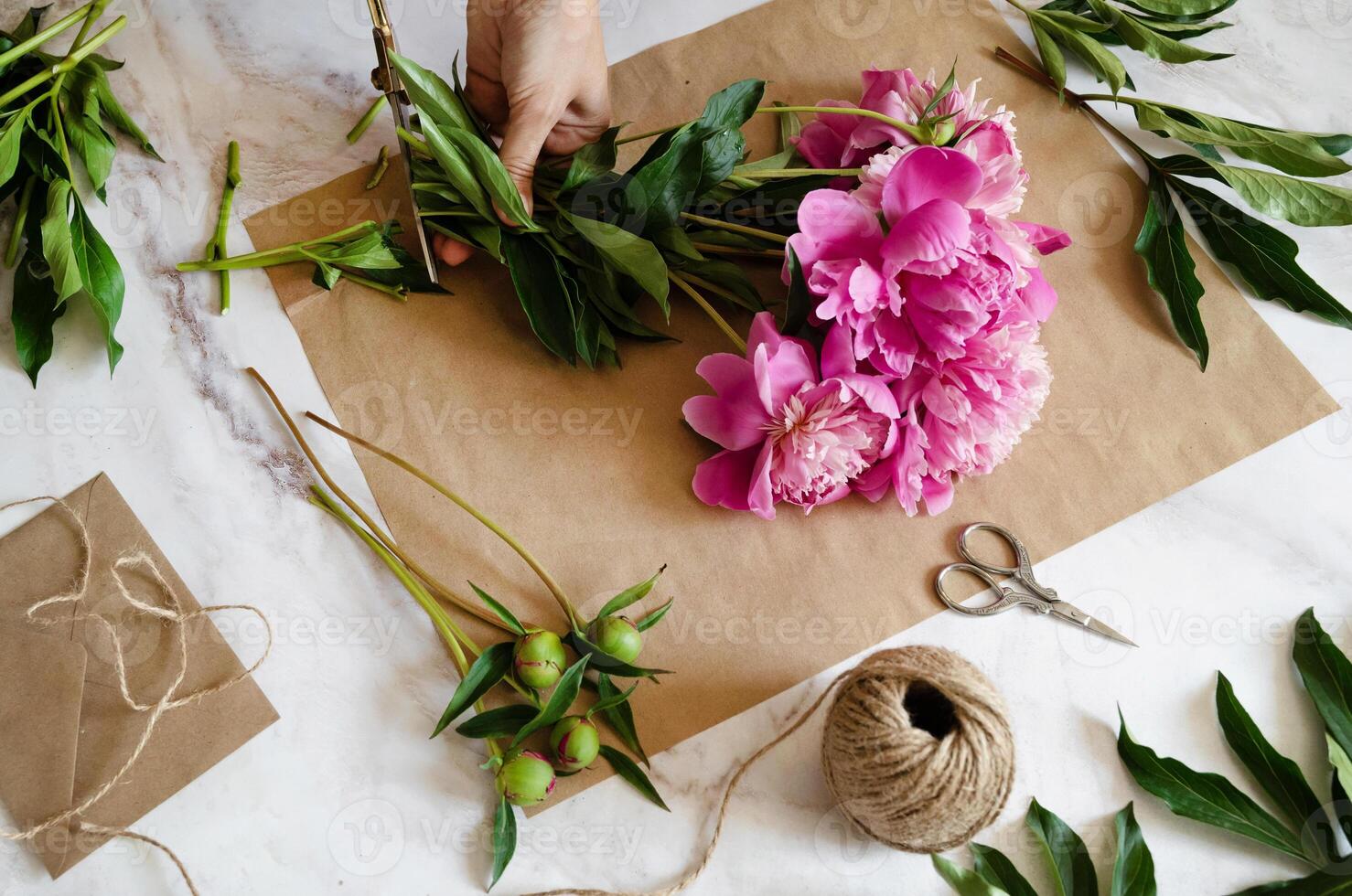 A floral composition in progress, featuring pink peonies, craft paper, scissors, and twine, suggesting a handmade and heartfelt creation. photo