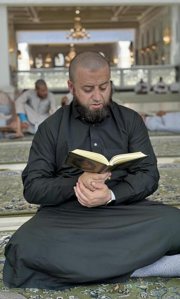 Mecca, Saudi Arabia - May 2, 2024. Young man, Hajj and Umrah pilgrim sitting and reading Quran in Masjidil Haram, Great Mosque in Makkah, Saudi Arabia. Hajj 2024. photo