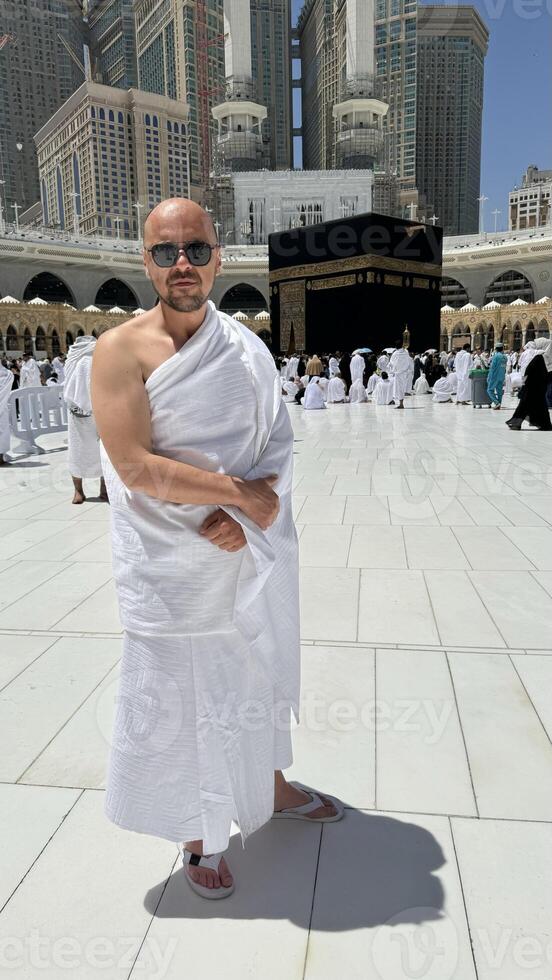 A bald man poses for a photo in front of the Kaaba at noon after performing Umrah at Masjid al-Haram, Mecca. A sacred Islamic pilgrimage in the heart of the holy mosque under the bright sunlight.