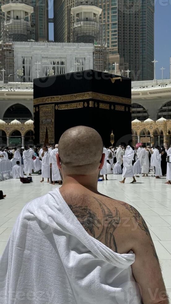 Former Sinner man who converted to Islam, with a large tattoo on his back and arm, praying with devotion in Makkah while gazing at the sacred Kaaba, embracing his new faith with deep spiritual photo