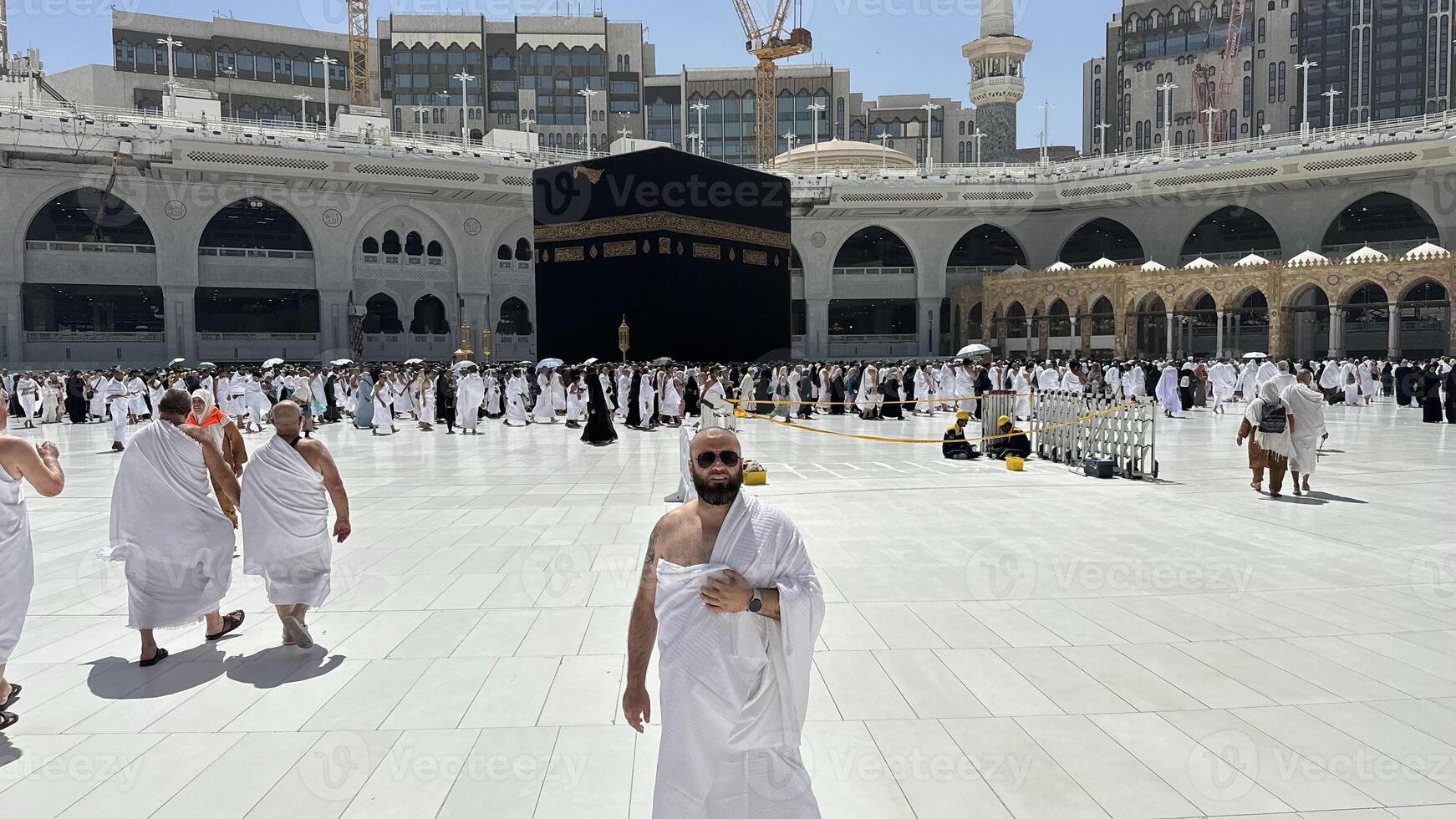 A bald man poses for a photo in front of the Kaaba at noon after performing Umrah at Masjid al-Haram, Mecca. A sacred Islamic pilgrimage in the heart of the holy mosque under the bright sunlight.