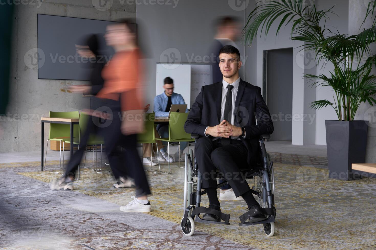 A businessman in a wheelchair navigating through a busy office, surrounded by his colleagues who are actively engaged in their work and collaboration photo
