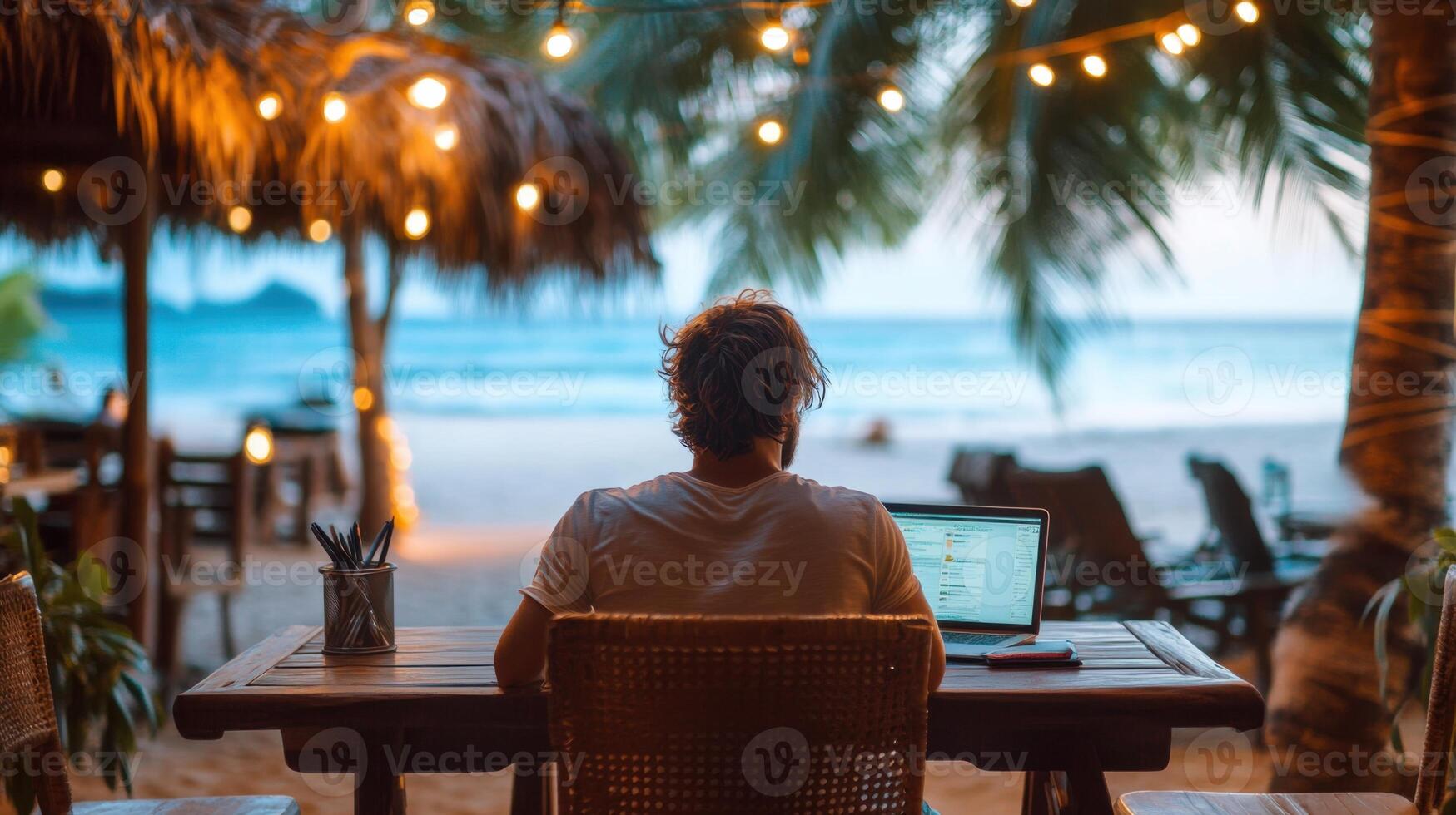 Freelancer Man at desk. Beach view. Working on computer. Lights above. Relaxing scene. Remote work. Tropical place. Outdoor office. Wooden table. photo