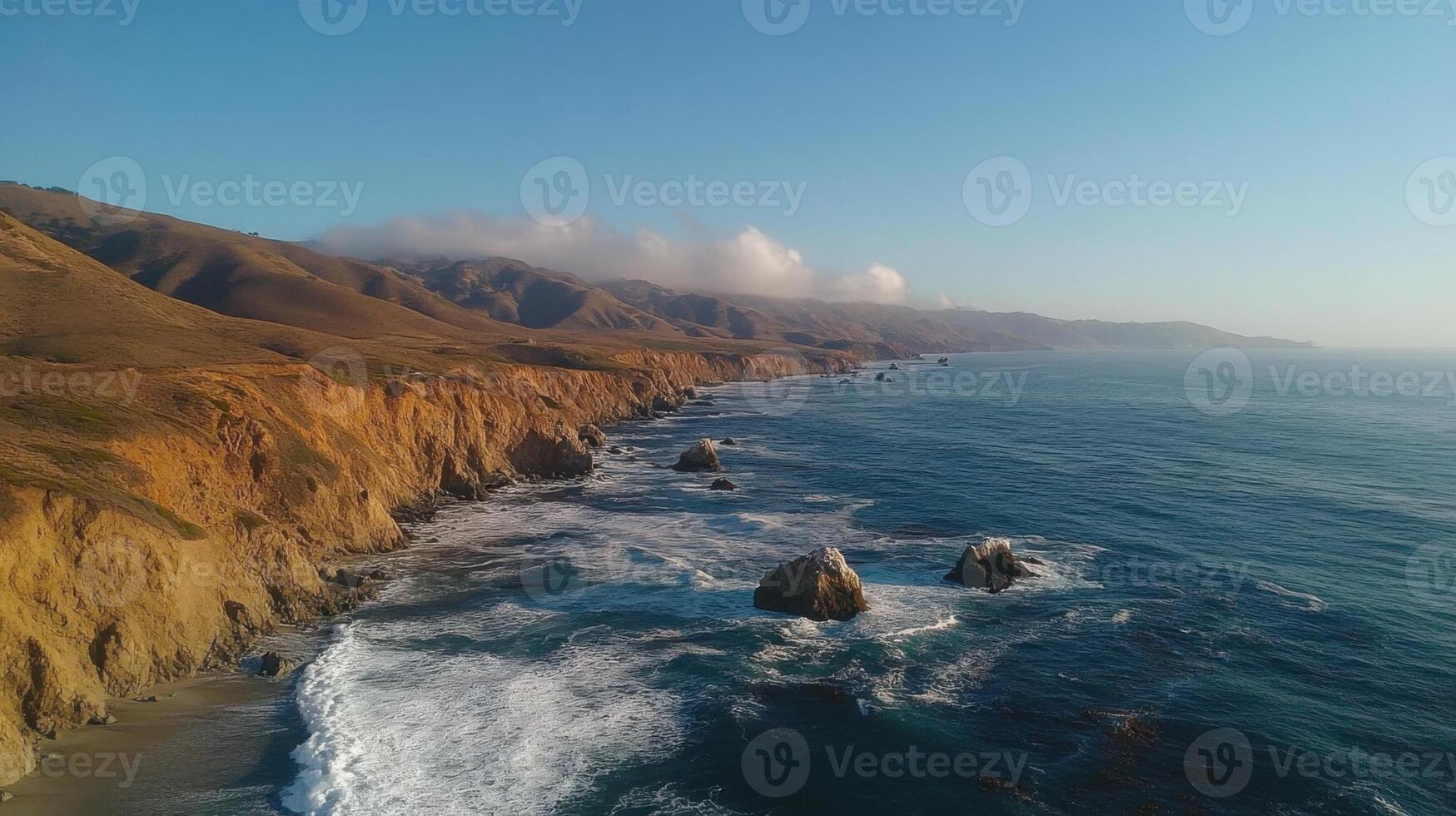 Ocean waves crash against rocky cliffs and beaches under a clear blue sky. Coastal hills and mountains rise in the background, shrouded by low clouds. photo