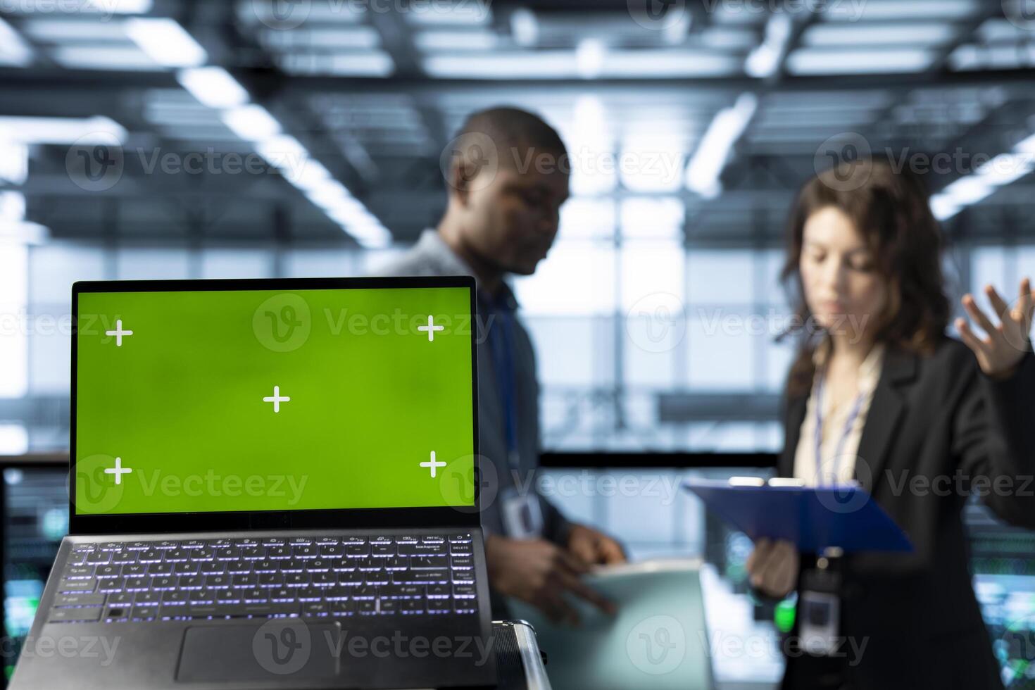 Green screen laptop in front of data center technicians in blurry background calibrating energy usage, inspecting UPS configurations, supporting sustainable infrastructure photo
