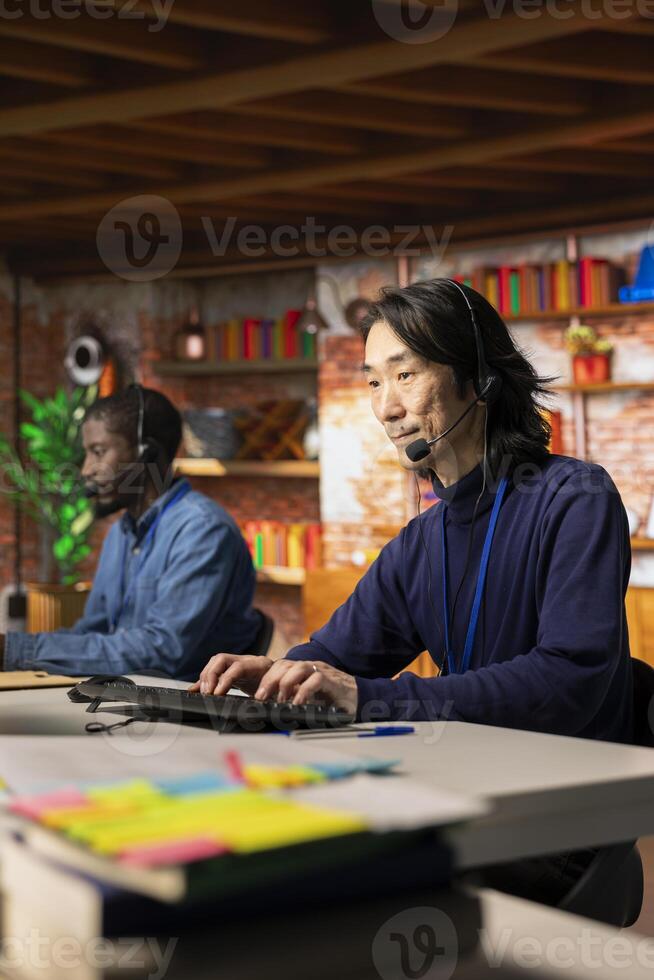 Experienced call center senior agent talking in headset mic, helping customers regain access to accounts and devices. Asian man in customer center assisting clients needing help resetting password photo