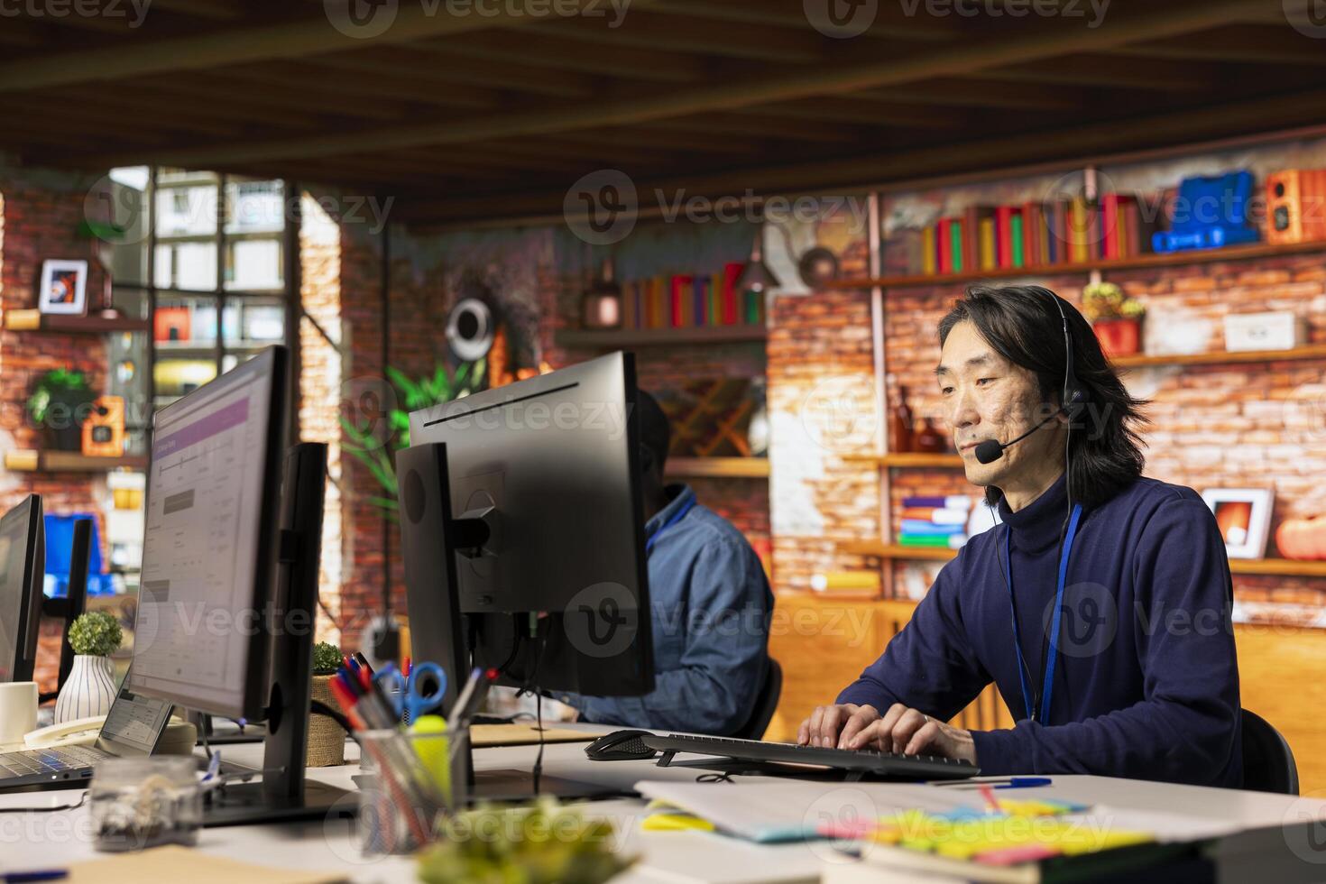Senior call center agent in office helps customers by answering inquiries, addressing issues. Experienced Asian man in customer support workspace in front of PC using headset mic to talk with clients photo