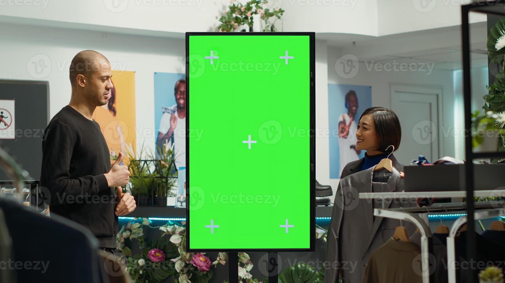 Asian retail worker recommends new designer items next to chroma key screen, suggesting colorful clothes for middle eastern man. Female assistant helping client with wardrobe essentials. photo