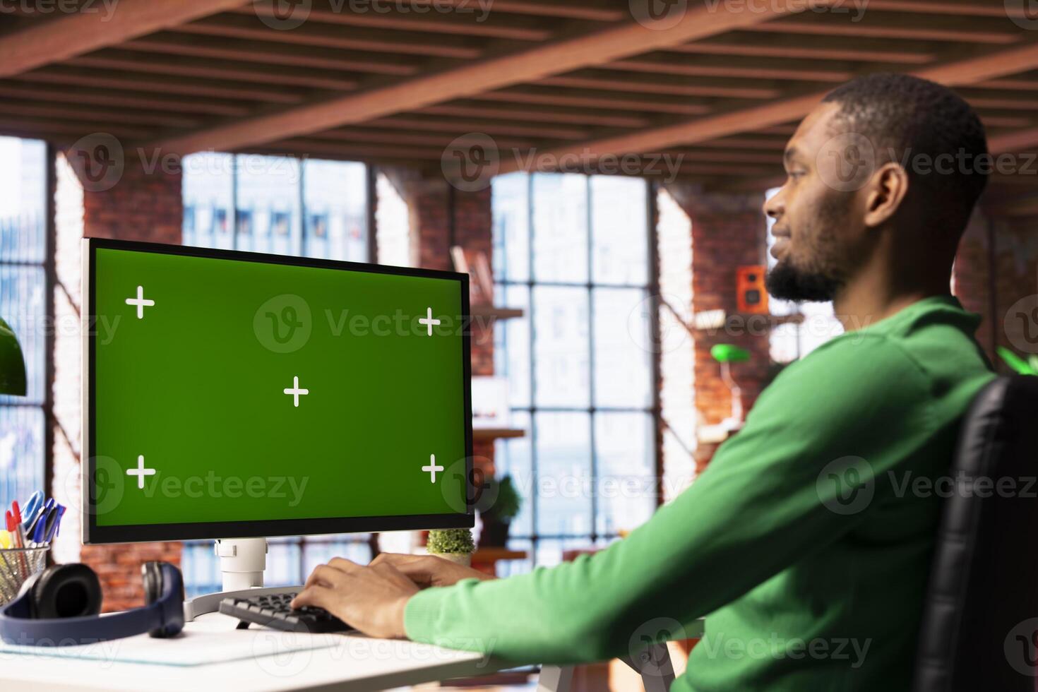 Smiling man enjoying leisure time in apartment, using green screen PC screen to text friends. Happy person talking with mates on social network website using chroma key computer display photo