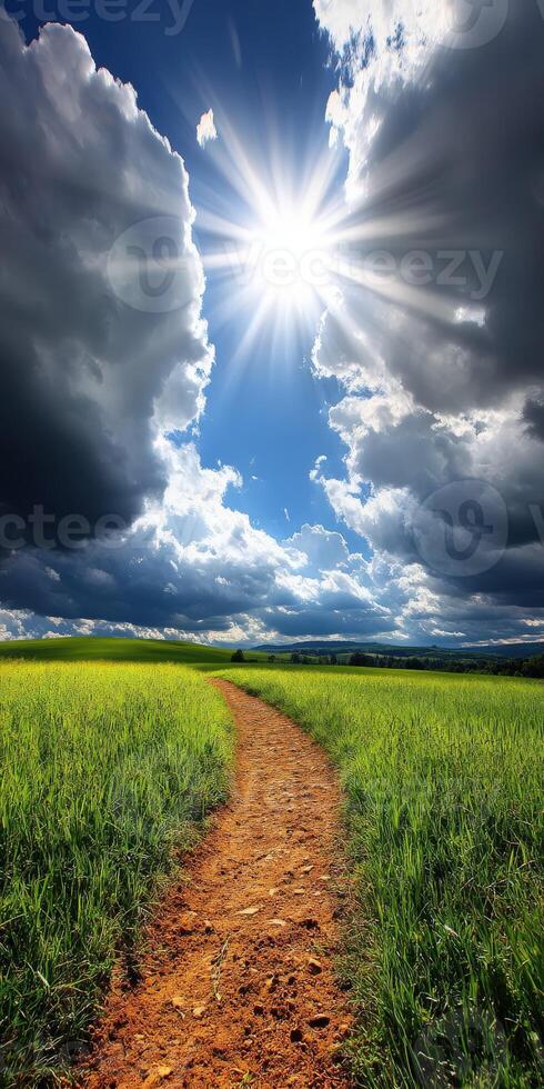 A dirt path in a grassy field with the sun shining through the clouds photo