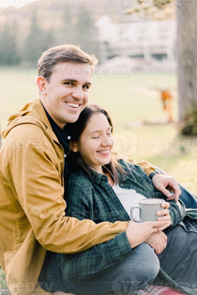 Smiling man hugging woman with mug sitting in autumn park photo