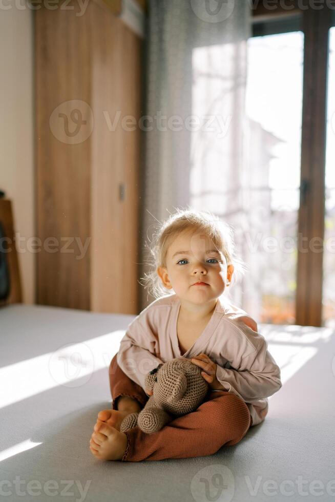 Little girl hugging a teddy bear while sitting on the bed in the bedroom and leaning forward photo