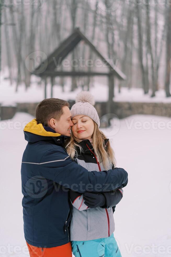 Smiling man hugging woman from behind, touching her temple with his nose in snowy park photo