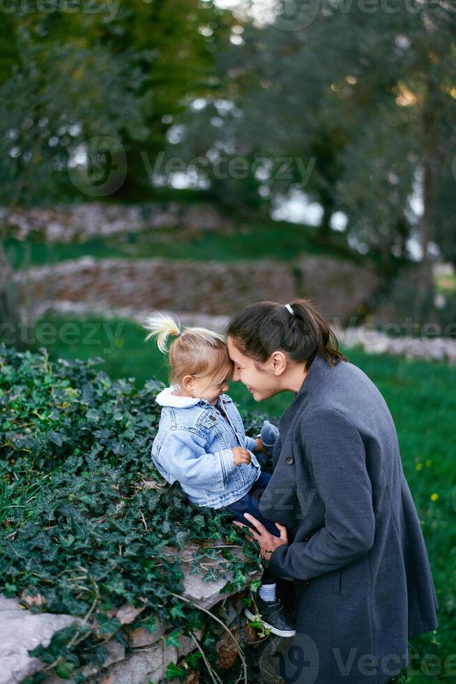 Mom and little girl touch their foreheads while sitting on a stone fence photo