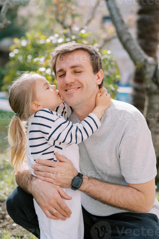 Little girl kisses her smiling dad on the cheek as he hugs her while squatting in the garden photo