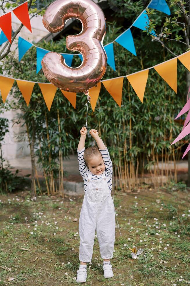 Little girl stands on a green lawn under colorful flags on a string and holds an inflatable number 3 photo