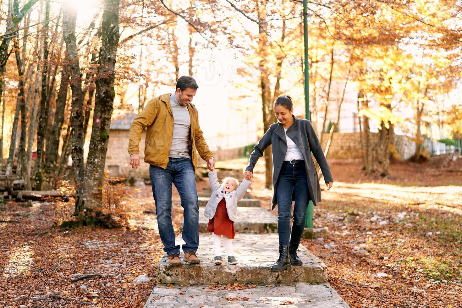 Little girl stands on a paved path in the park holding hands with mom and dad photo