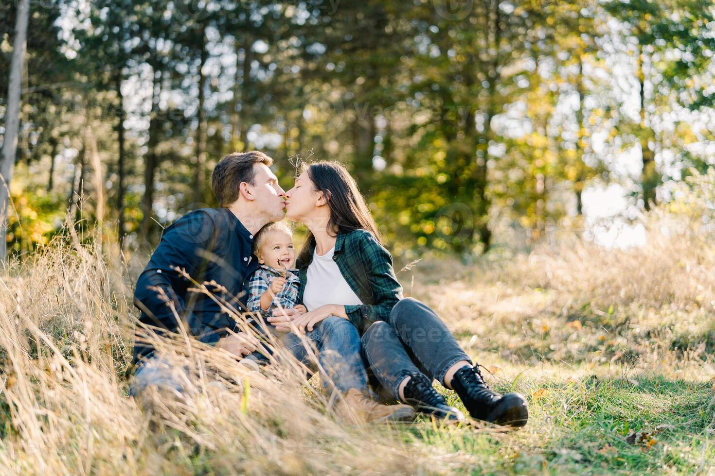 Dad and mom kiss while sitting with a little girl in a clearing near the forest photo